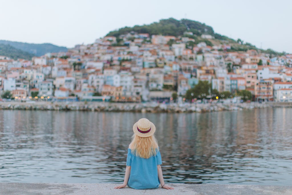 A woman in a blue dress sits on a pier, gazing at a picturesque coastal town.
